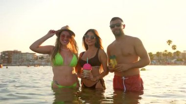 Neşeli turistler tropikal kokteyllerin ve ananasların keyfini çıkarırken altın deniz suyunda hareketli günbatımı anlarında kadeh tokuşturuyorlar.