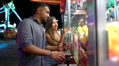 Neşeli genç çift, pençe turna oyunu oynuyor, gece vakti eğlence parkının ışıkları altında kahkaha ve heyecan paylaşıyorlar, birlikte romantik anları yakalıyorlar.