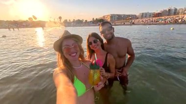 Üç arkadaş, iki kadın ve bir erkek, gün batımında denizde serinletici kokteyllerin tadını çıkarırken selfie çekiyorlar.