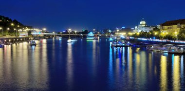 Vltava nehri Prag 'ın canlı şehir manzarasını yansıtıyor, sıcak bir yaz gecesinde gemiler zarifçe yelken açıyor, sakin bir atmosfer yaratıyor.