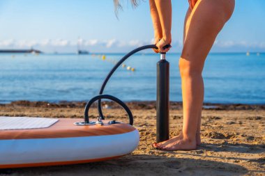 Kumsalda raket şişiren bir kadın yaz sörfü seansına hazırlanıyor. Sakin okyanus ve arka planda berrak gökyüzü