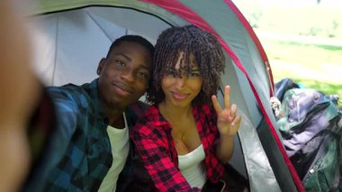 Mutlu genç çift selfie çekiyor, yaz tatili kaçamağı sırasında rahat bir kamp çadırında neşeli jestler yapıyor.