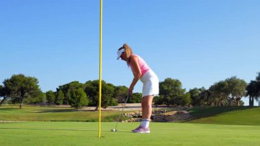 Profesyonel bayan golfçü, hassas bir şekilde batıyor. Başarılı bir şutu sevinçle kutluyor. Parlak yaz gökyüzü altında el değmemiş golf sahasında, destekleyici taşıyıcının yanında.