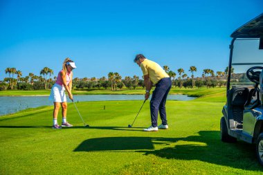 Baba-kız ikilisi güneşli bir günde golf oynuyor. Palmiye ağaçları ve açık mavi gökyüzü ile çevrili. Sahada kaliteli vakit geçiriyorlar.
