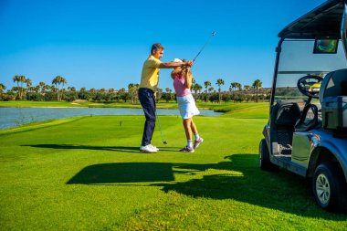Golf hocası, kadın golfçüye güneşli bir sahada rehberlik ediyor. Golf arabasının yanında sallanma tekniğini ve su tehlikesini geliştiriyor.