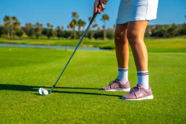 Dişi golfçü yoğun bir şekilde konsantre olurken yemyeşil golf sahasında güneşli ve parlak bir günde golf topuna vuruyor.
