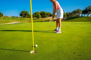 Bir kadın açık mavi gökyüzünün altındaki deliğe ustalıkla golf sahası kuruyor. Bereketli yeşil çevre huzurlu atmosferi güçlendirir.