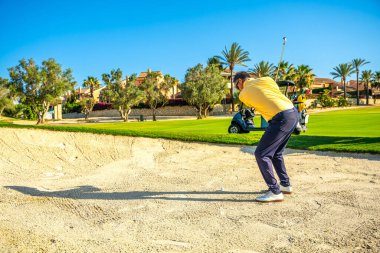 Golfçü, arka planda golf arabasıyla güneşli bir günde kum tuzağından topa vurmaya konsantre oluyor.