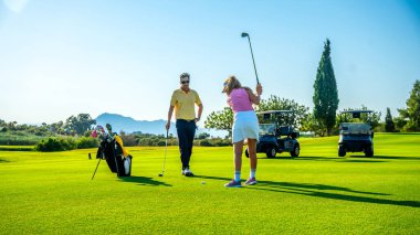 Golf hocası golf sahasında bayan golfçü çalıştırıyor. Güneşli bir günde arka planda golf arabaları var.