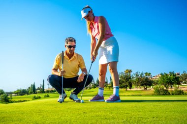 Golf hocası, açık mavi gökyüzü altında tekniğe odaklanan bir kadına golf sahasında rehberlik eder. Golf öğrenmek ve pratik yapmak için mükemmel bir gün.