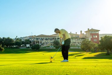 Golfçü bir yumruk sallıyor, topu canlı yeşil bir golf sahasında uçuruyor, arka planda lüks bir kulüp binası var.
