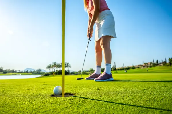 Güneşli bir günde topu deliğe sokan bayan golfçü, doğa ile çevrili çarpıcı bir golf sahasında eğleniyor.