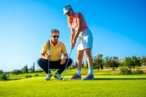 Golf hocası, açık mavi gökyüzü altında tekniğe odaklanan bir kadına golf sahasında rehberlik eder. Golf öğrenmek ve pratik yapmak için mükemmel bir gün.