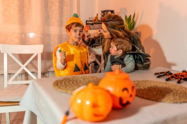 Anne çocuklarına cadılar bayramı makyajı yapıyor. Balkabağı gibi giyinmiş bir çocuk ve yarasa kostümü giymiş bir bebek.