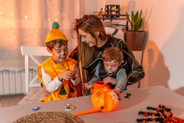 Cadılar Bayramı kostümü giyen anne ve çocuklar evde balkabağı kovası ve şekerlerle oynuyorlar.