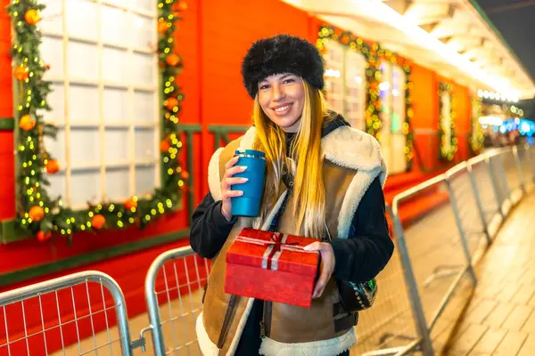 Genç bir kadın sıcak bir içecekten zevk alıyor ve bayram sezonunda bayramlık bir gece pazarında Noel hediyesi tutuyor.