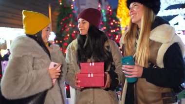 Noel hediyelerini paylaşan ve gece pazarında sıcak içeceklerin tadını çıkaran üç neşeli genç kadın.