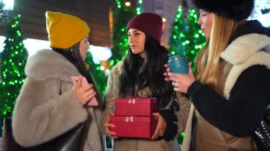 Üç neşeli kadın gece süslü bir şehir meydanında Noel hediyelerini ve sıcak içecekleri paylaşıyorlar.