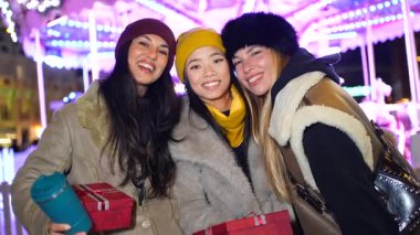 Üç neşeli arkadaş, kışlık giysiler içinde, bir noel pazarında parlak ışıklandırılmış bir atlıkarıncanın önünde kahkaha ve Noel hediyelerini paylaşıyorlar.