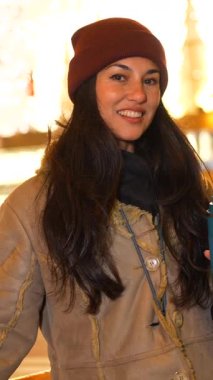 Kışlık kıyafetler giyen, elinde tekrar kullanılabilir bir kupa tutan, gülümseyen ve yılbaşı gecelerinin tadını çıkaran genç bir kadın.