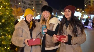 Üç neşeli kadın Noel hediyelerini tutuyor ve süslü bir Noel pazarında sıcak içeceklerin tadını çıkarıyor.