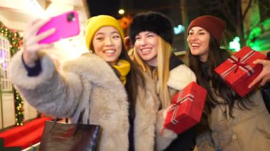 Süslü bir şehirde Noel hediyeleriyle selfie çeken üç neşeli genç kadın.