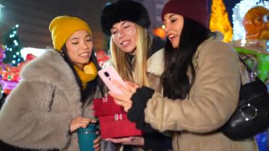Şehirde Noel gecesi eğlenen üç neşeli kadın hediye alışverişi yapıyor ve akıllı telefonuyla fotoğraf çekiyor.
