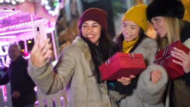 Arkadaşlar gülüyor, renkli Noel hediyeleriyle selfie çekiyor, kış tatili akşamları aydınlık gece pazarında neşeli anlar yakalıyorlar.