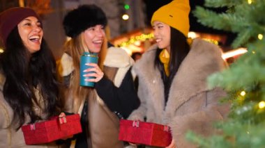 Üç neşeli kadın Noel hediyelerini değiş tokuş ediyor ve gece süslü bir Noel pazarında sıcak içecekler içiyor.