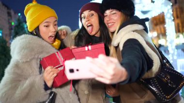 Üç neşeli genç kadın neşeli anıları yakalıyor, paketlenmiş noel hediyeleriyle poz veriyorlar, parıldayan şehir yılbaşı ışıklarının arasında selfie çekiyorlar.