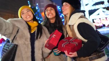 Genç kadınlar Noel hediyeleriyle selfie çekiyorlar. Aydınlatılmış gece pazarında Noel süsleriyle neşeli anları yakalıyorlar.