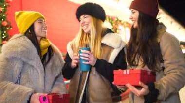 Noel hediyelerini değiş tokuş eden ve gece pazarında sıcak içeceklerin tadını çıkaran üç neşeli genç kadın.