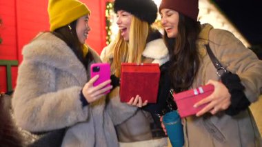 Noel hediyelerini değiş tokuş eden ve süslü bir gece marketinde fotoğraf çeken üç neşeli genç kadın.