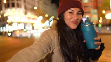 Neşeli genç kadın sıcak bir içecek içiyor. Parlayan yılbaşı ışıkları ve bulanık şehir gece manzarası.