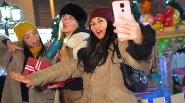 Mutlu bayan arkadaşlar gece süslü bir şehirde yılbaşı hediyeleriyle selfie çekiyorlar.