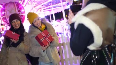 Mutlu arkadaşlar fotoğraf çekerler ve gece bir atlıkarınca önünde Noel hediyelerini paylaşırlar.