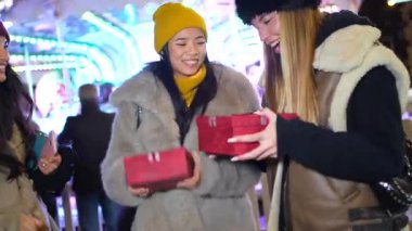 Şenlik neşesini paylaşan genç kadınlar, gece vakti kentsel kış kutlamaları sırasında aydınlatılmış pazar tezgahlarının yakınında paketlenmiş noel hediyelerini paylaşıyorlar.