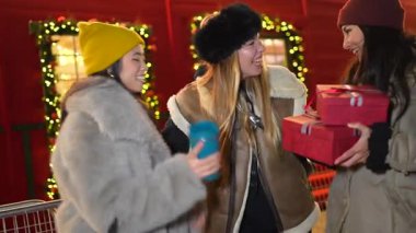 Neşeli genç kadınlar parıldayan şehir ışıkları altında Noel hediyelerini paylaşıyorlar, kış akşamları dostluk ve tatil neşesi yayıyorlar.