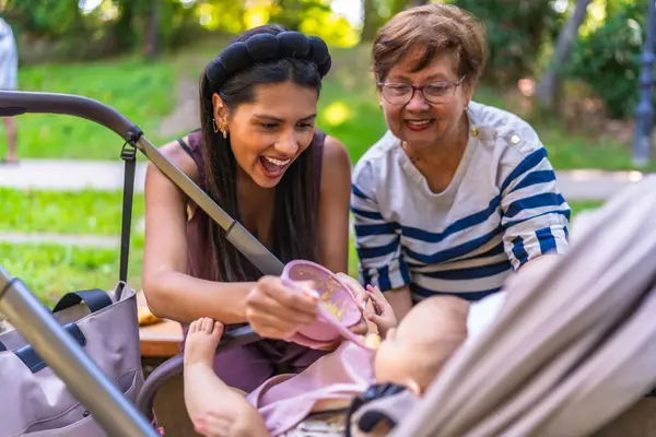 Latin anne bebeğini pembe bir kaseden katı gıdayla besliyor. Büyükannesi de parkta yardımcı oluyor.