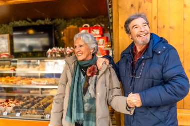Kış tatilinde geleneksel bir Noel pazarını ziyaret ederken el ele tutuşan ve gülümseyen neşeli yaşlı çift.