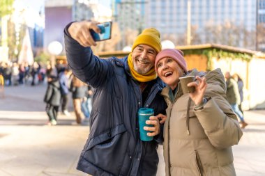 Kış şehrindeki bir Noel pazarında sıcak içeceklerin tadını çıkarırken akıllı telefonuyla selfie çeken gülümseyen yaşlı çift.