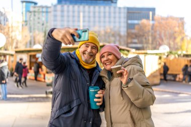 Gülümseyen son sınıf çifti kentteki bir Noel pazarında selfie çekiyor ve sıcak içecekler içiyor.