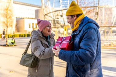 Yaşlı bir adam, güneşli bir kış parkında mutlu bir şekilde gülümseyen bir kadına Noel hediyesi veriyor, aşkı ve birlikteliği kutluyor.