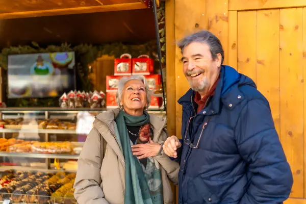 Kış şehrindeki bir Noel pazarında gülümseyen ve eğlenen neşeli yaşlı çift.
