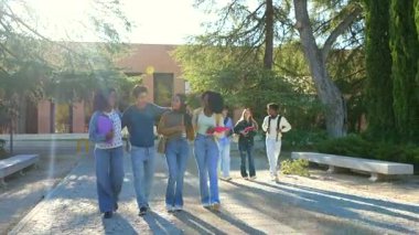 Üniversite kampüsünde birlikte yürüyüp konuşan neşeli çoklu etnik arkadaş grubu, ellerinde kitaplarla dersten sonra güneşli bir günün tadını çıkarıyorlar, eğitimi ve gençlik arkadaşlığını temsil ediyorlar.