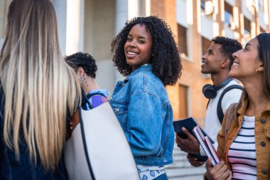 Bir grup çok ırklı üniversite öğrencisi kampüste birlikte yürüyor, gülümsüyor ve etrafa bakınıyor, akademik bir ortamda gençliği, eğitimi ve arkadaşlığı temsil ediyorlar.