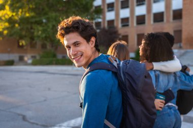 Üniversite öğrencisi sırt çantasıyla kameraya gülümsüyor, kampüste arkadaşlarıyla yürüyor, üniversite deneyimi sırasında mutluluk ve bağlantı gösteriyor.
