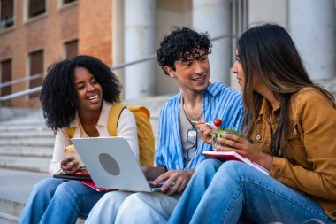 Çok ırklı üniversite öğrencilerinden oluşan bir grup açık hava basamaklarında oturuyor, hafif bir yemek paylaşıyor, gülüyor ve sosyal bir mola sırasında dizüstü bilgisayar kullanarak iletişim kuruyor.