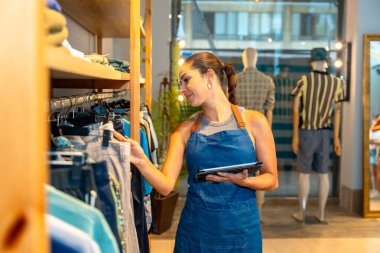 Genç satış asistanı bir moda mağazasında elbise envanterini kontrol ederken dijital tablet kullanıyor.
