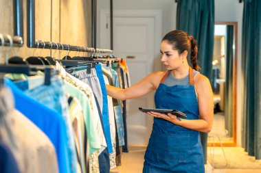 Genç satış asistanı, şık bir perakende mağazasında askılarda asılı olan giysi parçalarını kontrol etmek için dijital tablet kullanıyor ve envanteri verimli bir şekilde yönetiyor.